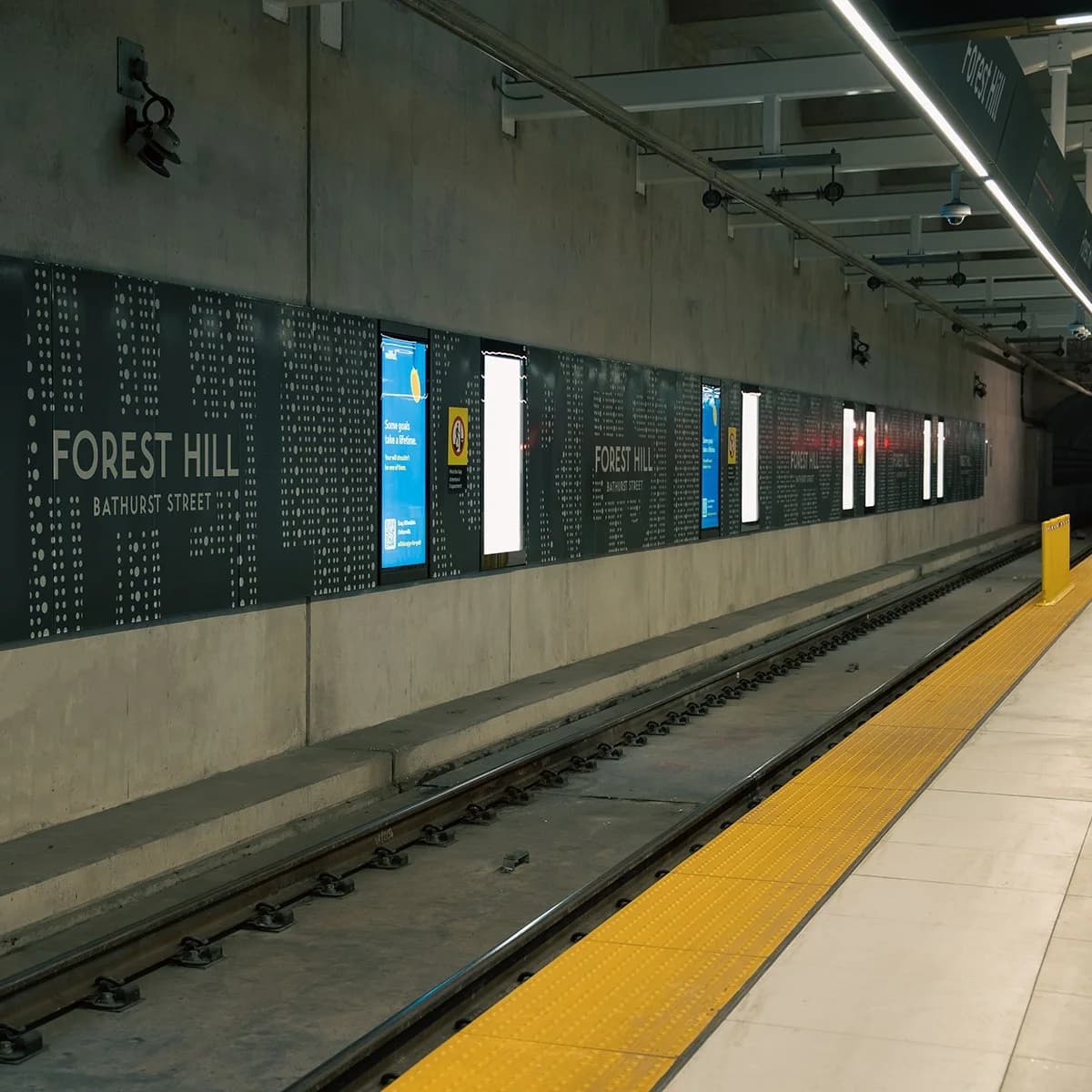 Forest Hill (Bathurst Street) metro istasyonunda peron: duvar boyunca noktalı paneller ve içten aydınlatmalı reklam ekranları, sarı uyarı şeridi ve raylar