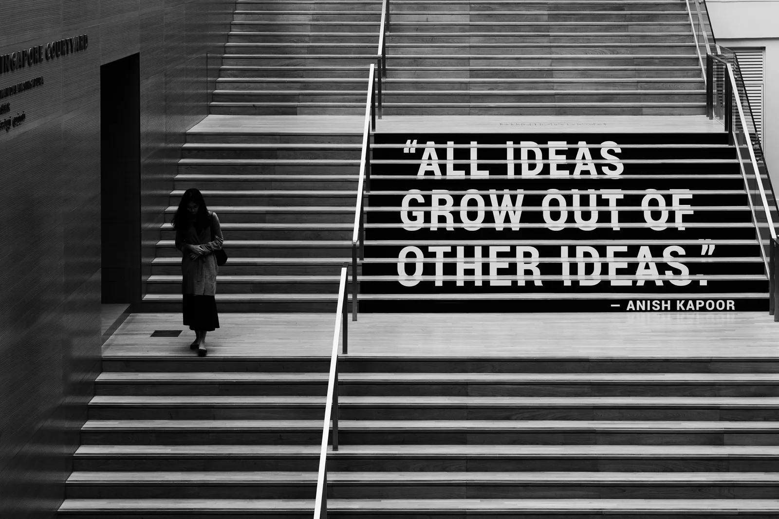 Siyah beyaz fotoğraf: geniş bir ahşap merdivenin basamaklarına yazılmış "All ideas grow out of other ideas" (Anish Kapoor) sözü; solda merdivenden inen bir kişi